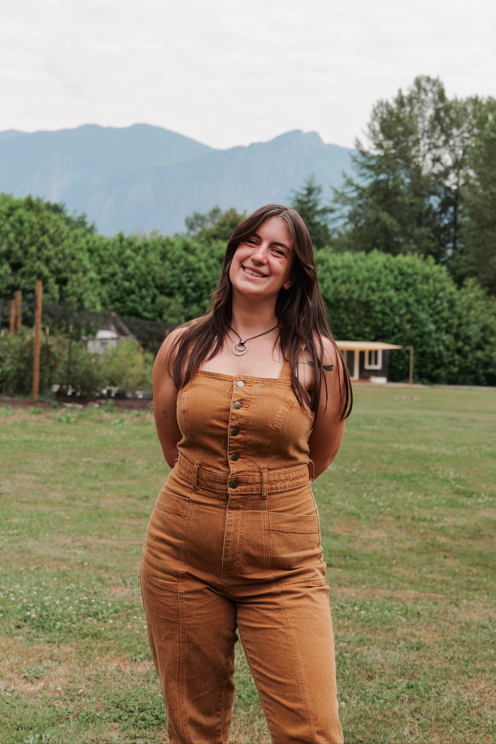 Woman in a mustard yellow jumpsuit standing in a grassy field with trees and mountains in the background
