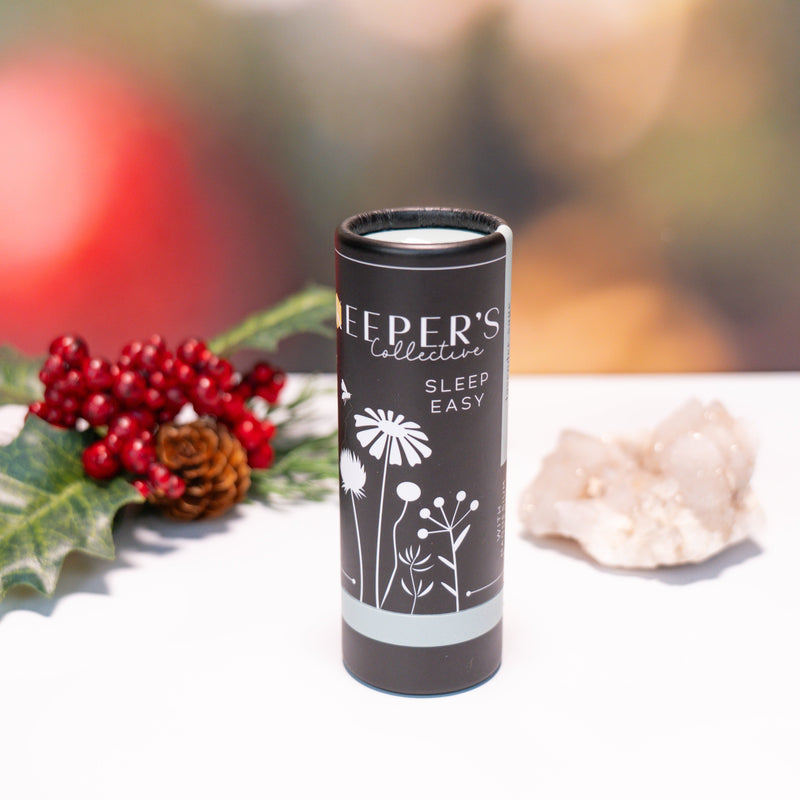 Candle in a black container with 'Sleep Easy' text, surrounded by holly and pine cones on a white surface.